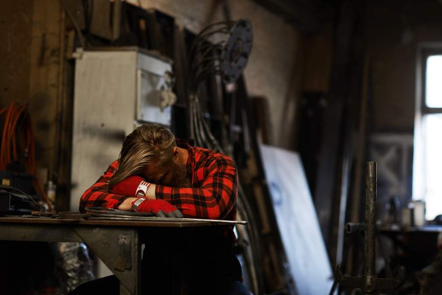 Müder und überarbeiteter Mitarbeitnehmer, der mit dem Kopf auf den Armen an einem Tisch in der Arbeitsumgebung schläft, braucht etwas Ruhe, um Müdigkeit zu reduzieren.