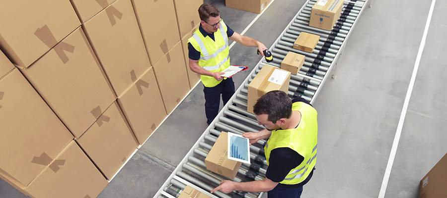 Arbeiter in Warnwesten stehen am Fließband und sortieren Pakete mit Tablets und Handscanner in einem optimierten Arbeitsablauf